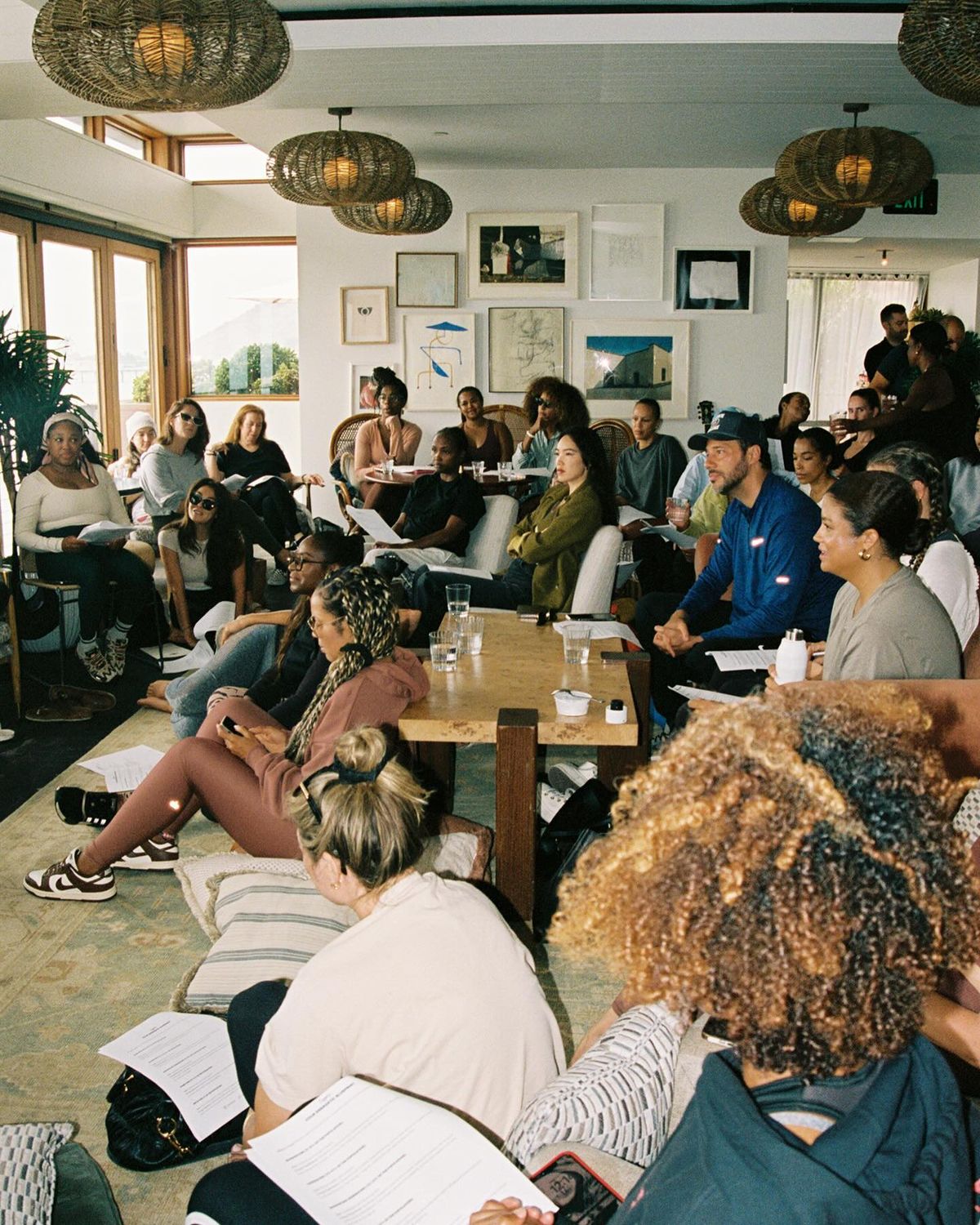 A mid-sized group of multi-racial people sitting in a room, some on pillows on the floor and other on couches and chairs across the room.