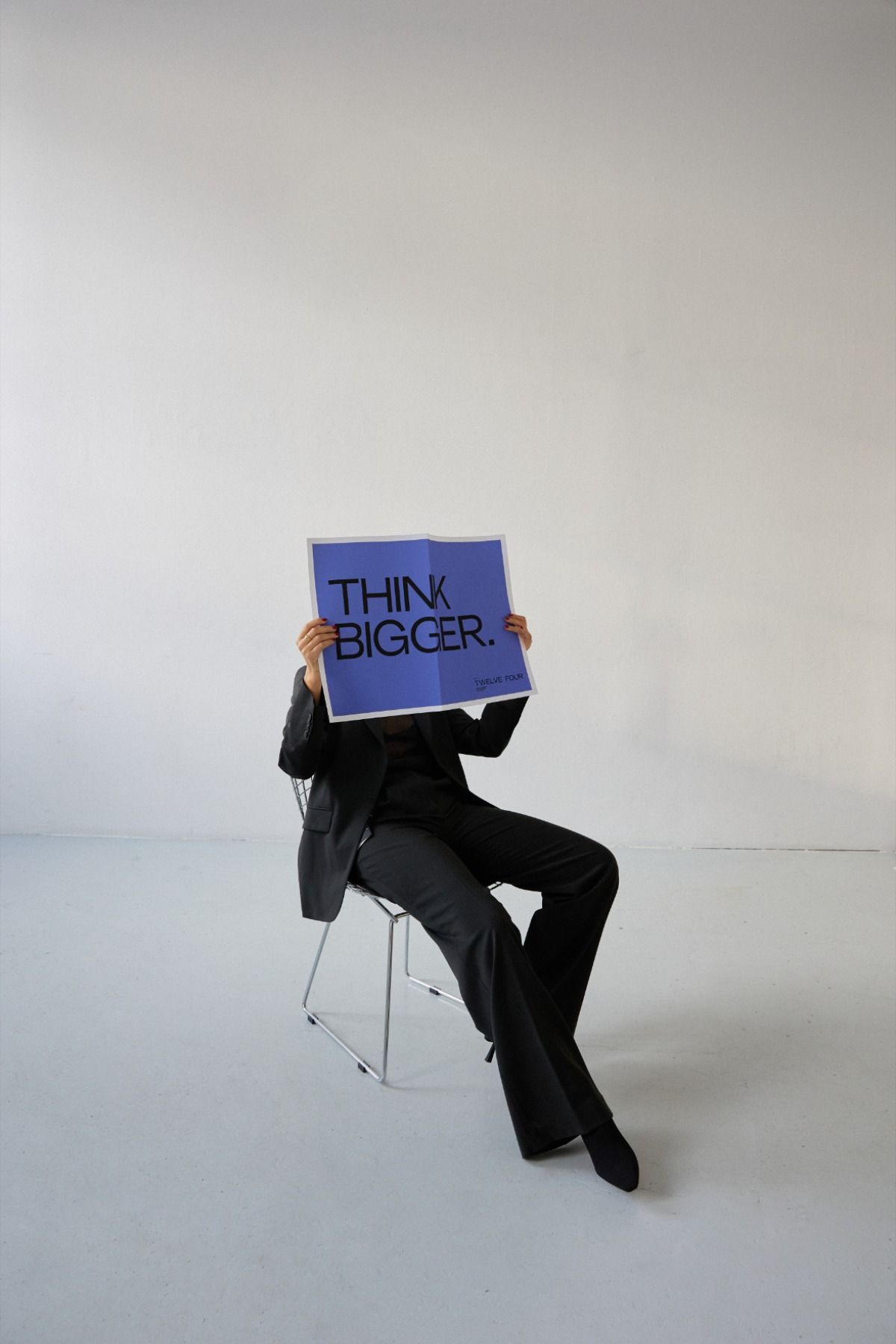 Person sitting in a photo studio in a black suit, holding a placard written "Think Bigger"